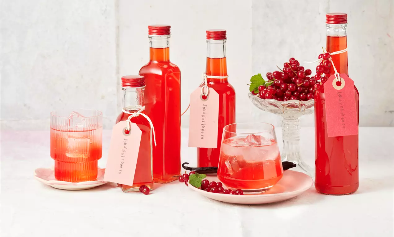 Roter Johannisbeerlikör in klaren Flaschen mit roten Schraubverschlüssen, beschrifteten Anhängern und Eisgläsern, dekoriert mit roten Johannisbeeren