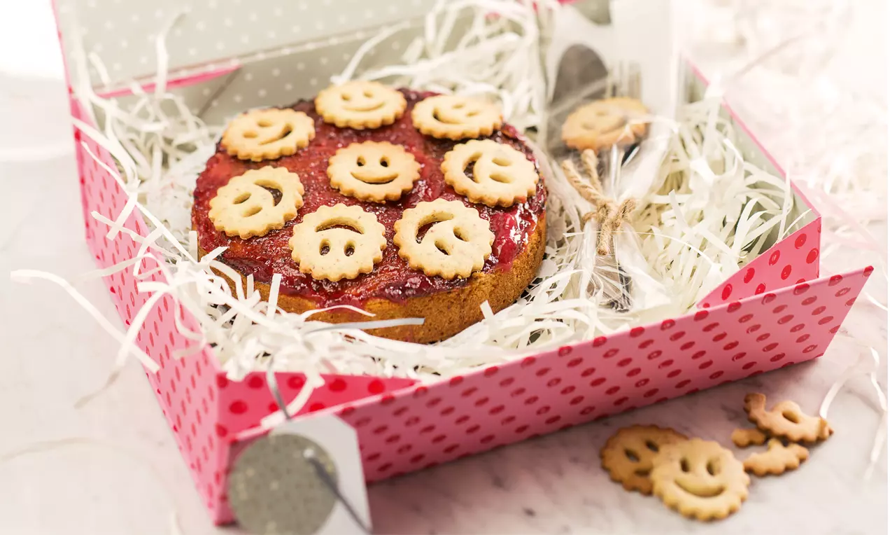Runde Linzer Torte mit roter Marmelade und Mürbeteigdekor in Smiley-Form, präsentiert in einer rot gepunkteten Schachtel mit Holzwolle