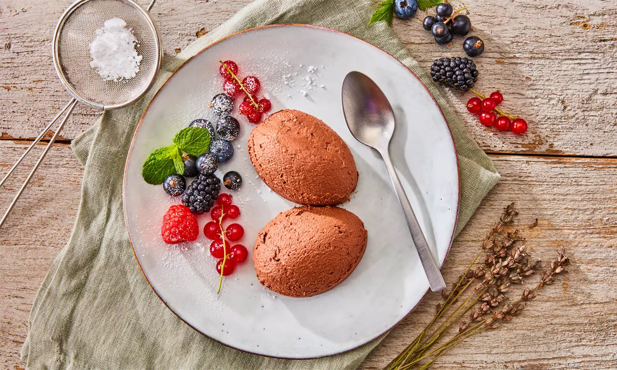 Schokoladenbraune Mousse-Kleckse mit cremiger Textur auf weißem Teller, umrahmt von frischen Beeren, Minzblättern und silbernem Löffel