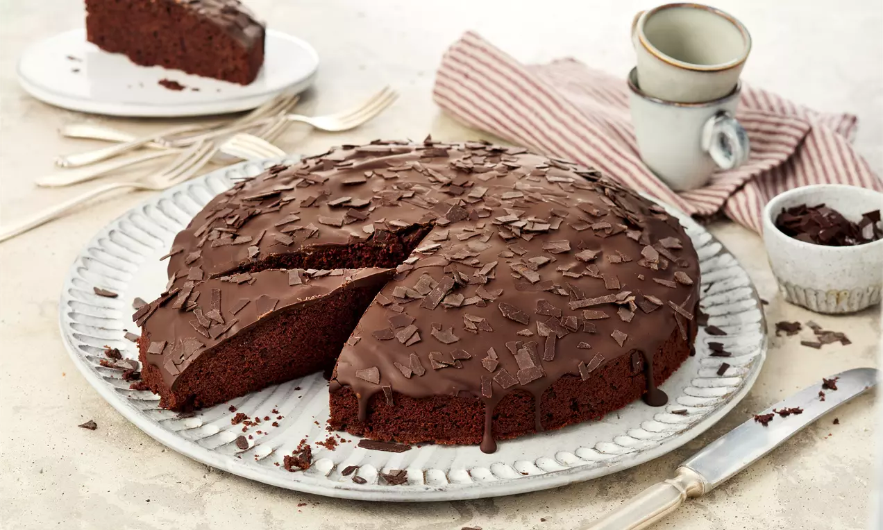 Veganer Schokokuchen auf einer weißen Platte, überzogen mit glänzender Schokoglasur und bestreut mit Schokoraspeln. Ein Stück ist herausgeschnitten. Daneben Besteck, Tassen und Serviette.