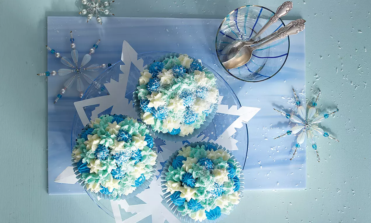 Eisblaue Cupcakes mit floraler Creme-Dekoration in Blau und Weiß auf einem gläsernen Teller mit Schneeflocken-Motiv, umgeben von zartem Winterdekor