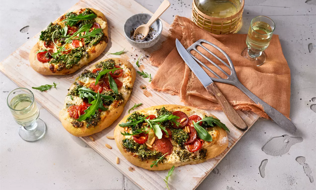 Drei goldbraune Rahmfladen mit cremigem Belag aus Bärlauchpesto, frischen Tomatenscheiben und grünem Rucola auf Holzbrett angerichtet