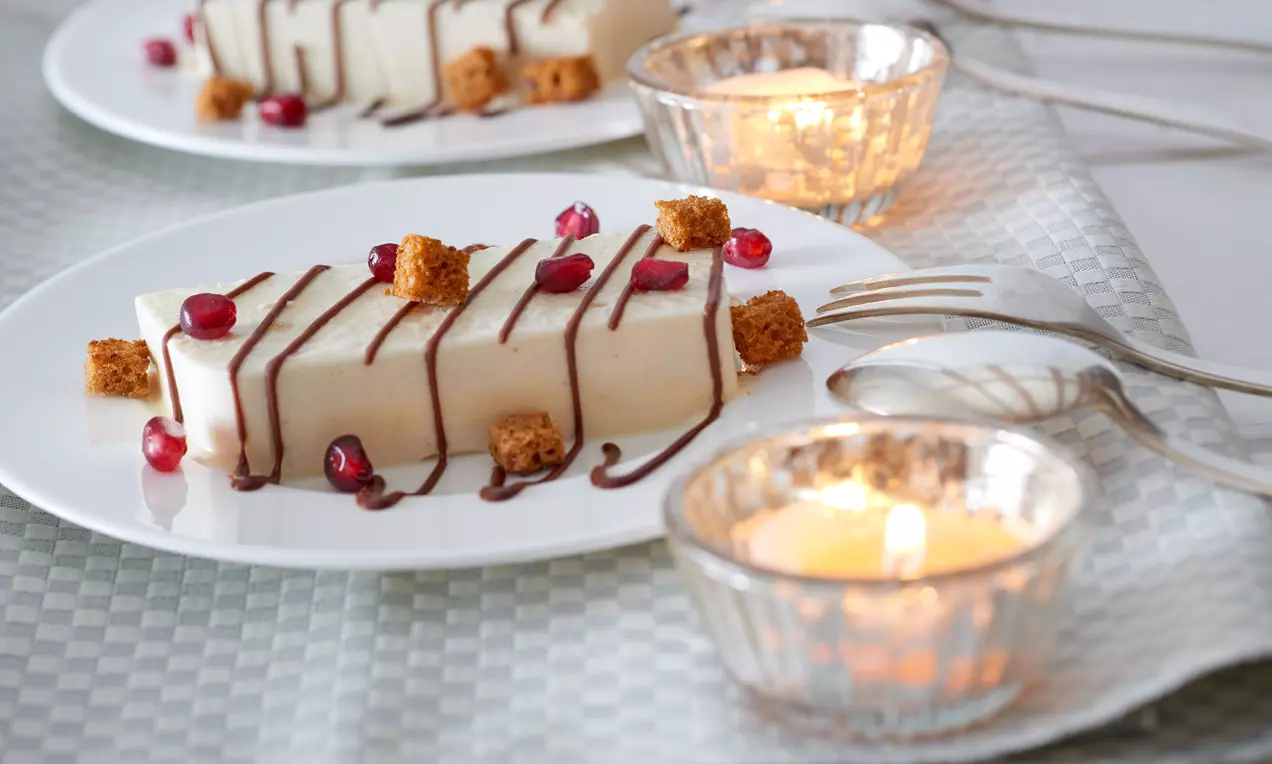 Lebkuchen-Parfait in heller Rechteckform mit Schokoladensoße, Granatapfelkernen und Lebkuchenstücken auf weißem Teller mit Kerzendekoration