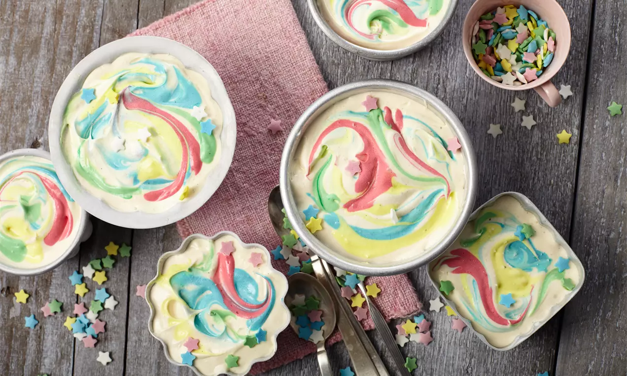 Buntes Eis mit geschwungenen Mustern aus Rosa, Blau, Gelb und Grün in verschiedenen Schalen, umgeben von bunten Sternstreuseln auf Holzoptik