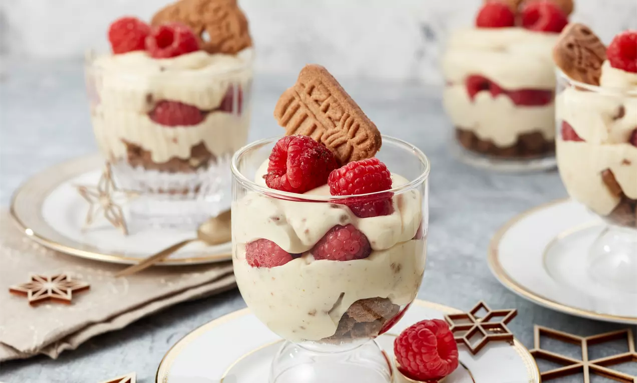Schichtdessert aus hellbeiger Creme, roten Himbeeren und braunem Spekulatius-Keks in einem gläsernen Dessertbecher auf weiß-goldenem Teller