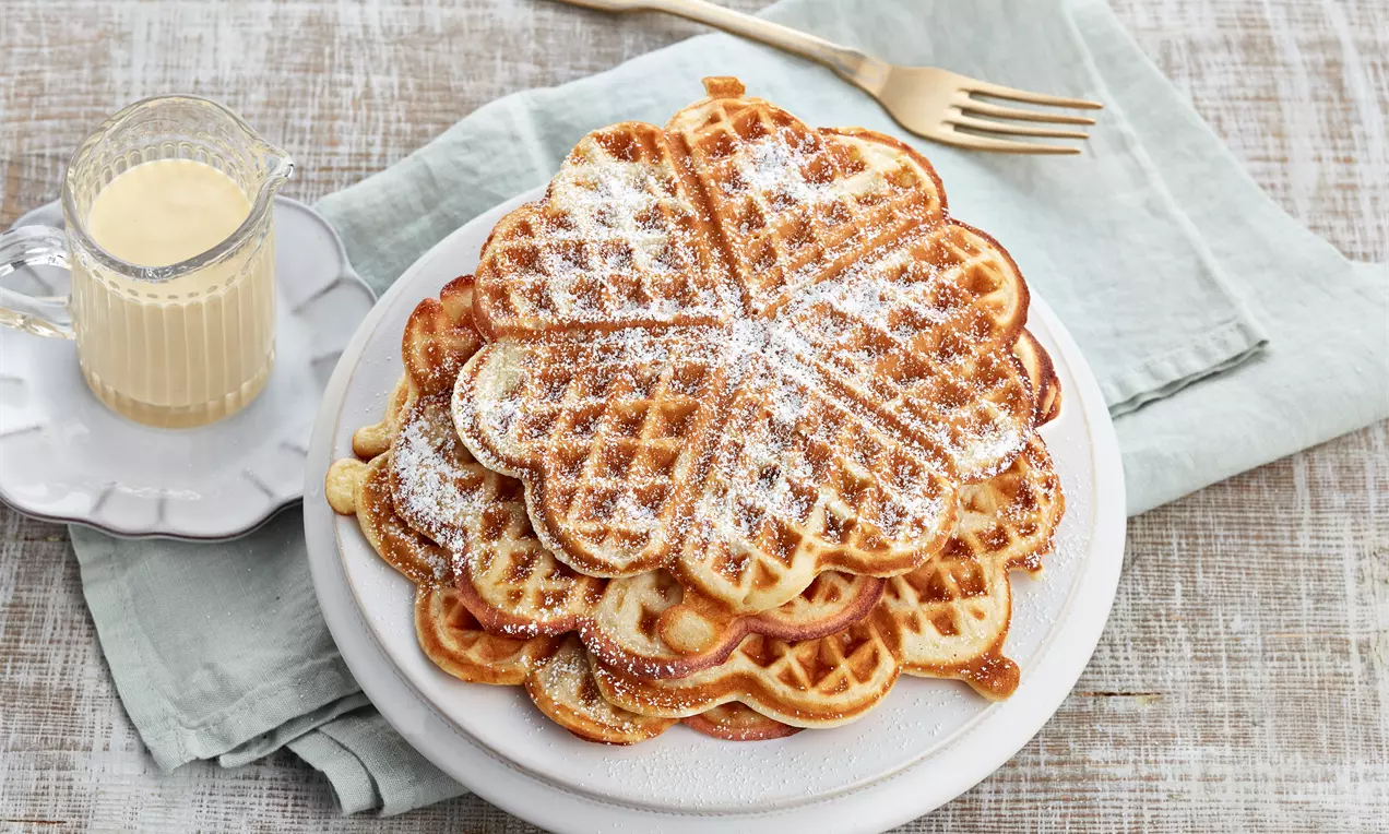 Goldbraune, herzförmige Apfel-Waffeln mit Puderzucker bestäubt auf einem weißen Teller, daneben helle Vanillesauce in einer kleinen Glaskanne