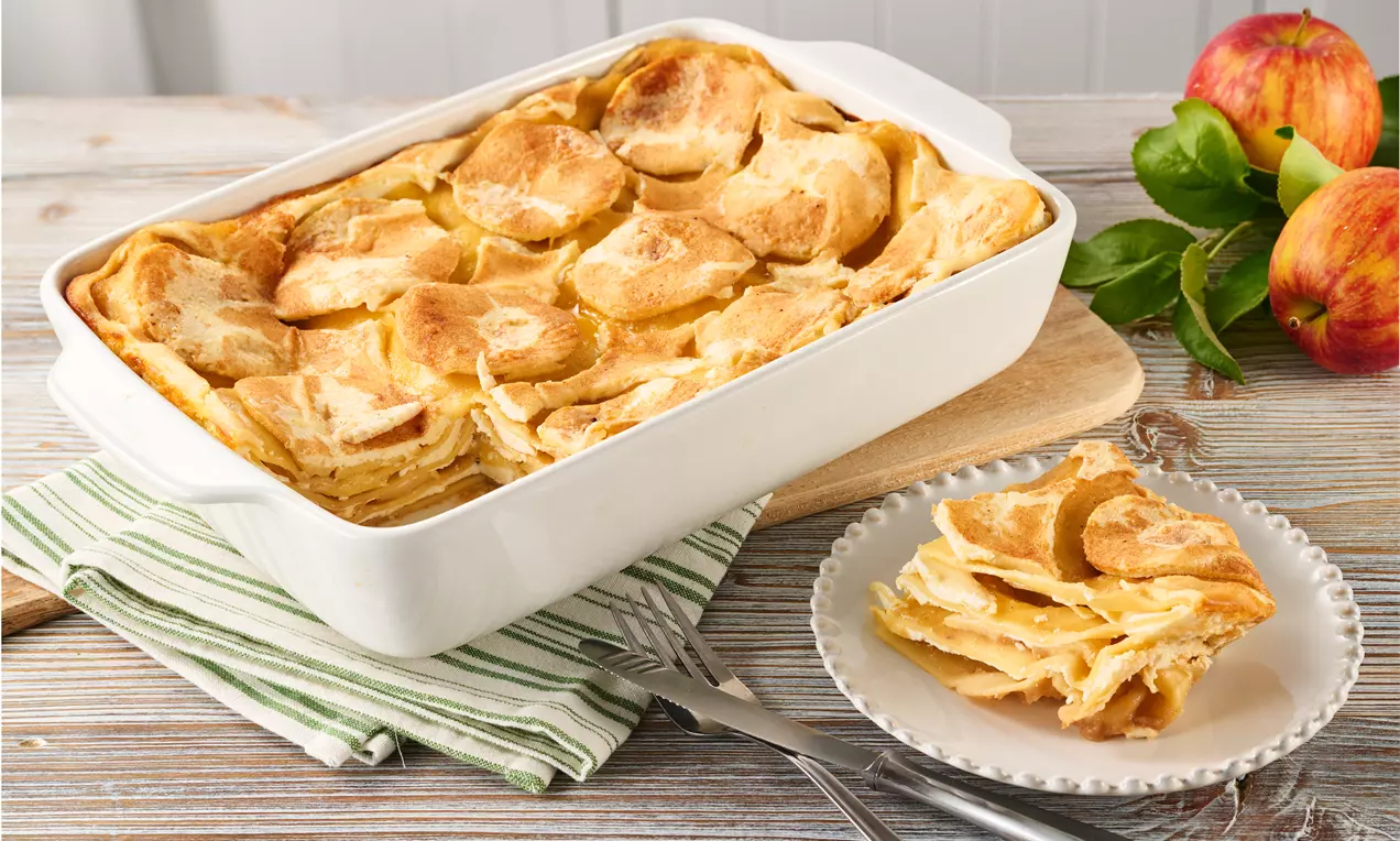 Goldbraune Apfellasagne mit dünnen, geschichteten Teig- und Apfelstücken in einer weißen Auflaufform, angerichtet auf Holzbrett mit Teller.