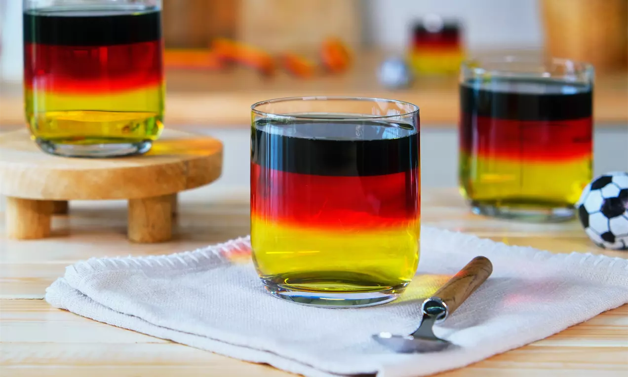 Farbschichtendes Dessert in Deutschlandflaggenfarben, gelb unten, rot in der Mitte, schwarz oben, in Gläsern auf einem hellen Tuch mit Löffel