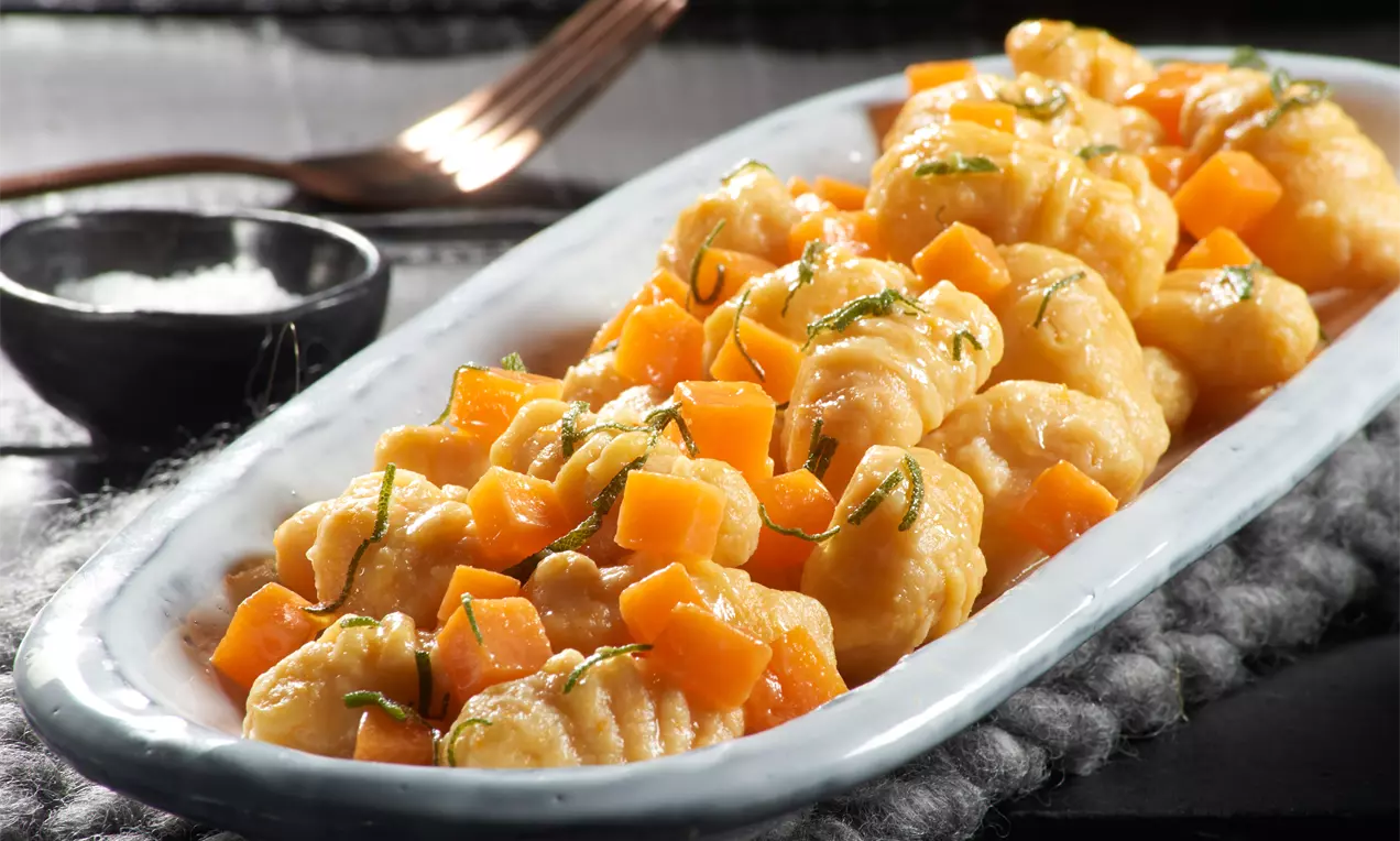 Kürbis-Gnocchi mit goldgelber Salbeibutter und orangefarbenen Kürbiswürfeln auf länglicher weißer Platte mit dunkelgrauer Unterlage