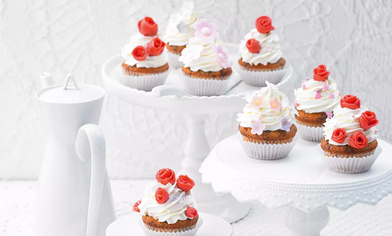 Kleine Cupcakes mit weißer Sahnehaube, roten Zuckerröschen und feinen Zuckerblüten, arrangiert auf weißen Etageren vor Spitzenhintergrund