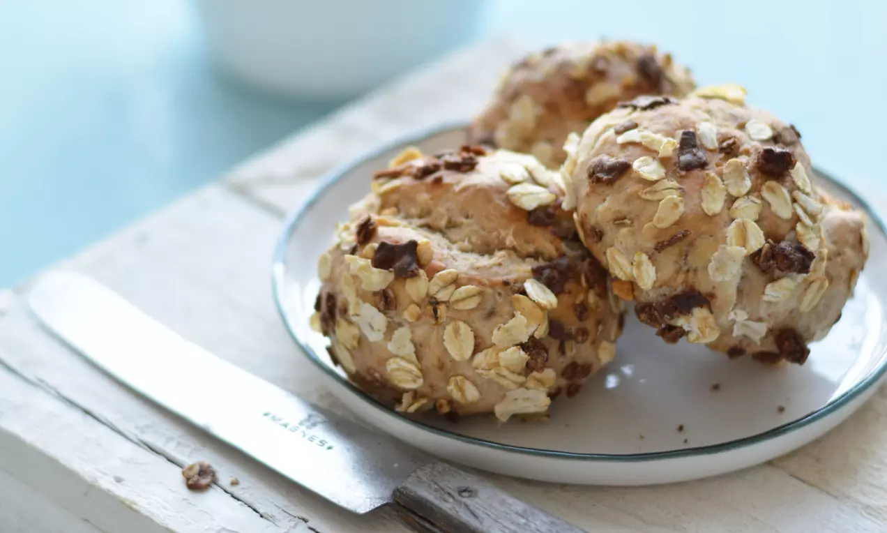 Kleine, goldbraune Müsli-Brötchen mit Haferflocken und Schokostückchen auf einem weißen Teller, daneben ein Messer mit Holzgriff