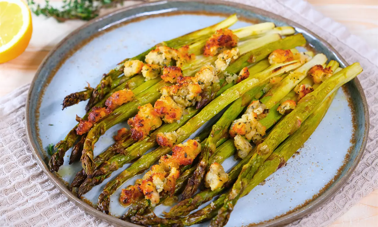 Goldbraune Spargelstangen mit knuspriger Kruste auf hellblauem Teller, umgeben von Kräutern und einer halbierten Zitrone