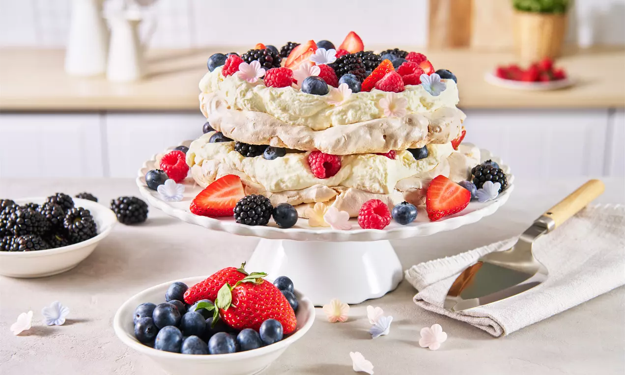 Zweistöckige Pavlova-Torte mit knuspriger, weißer Baiserschicht, Sahne, frischen Beeren und bunten Zuckerdekorationen auf einer Tortenplatte