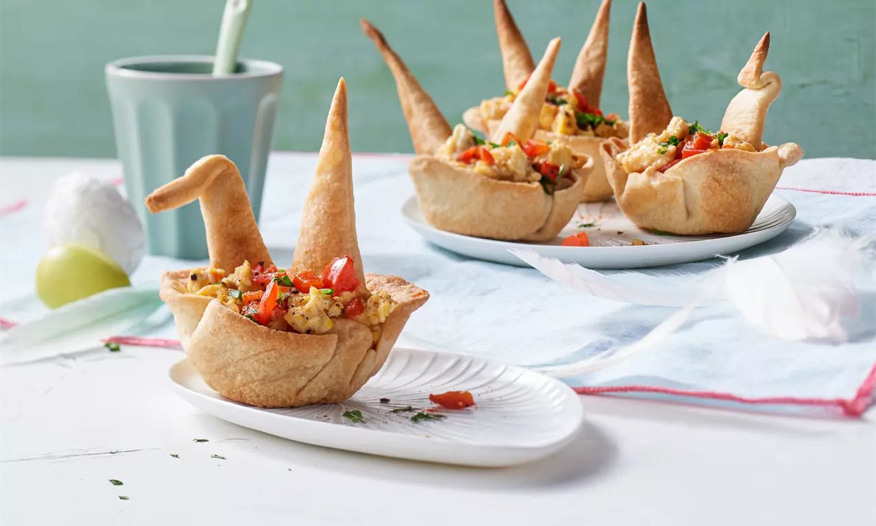 Knusprige Teigschalen in Form von Hasen mit goldgelbem Rührei, frischen Tomaten und Kräutern, auf weißem Geschirr mit hellblauer Dekoration