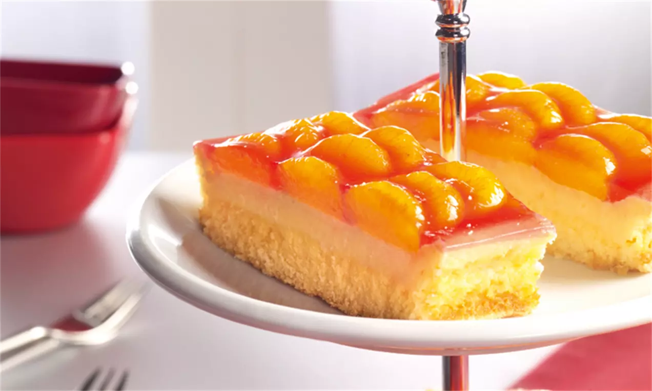 Mandarinen-Käsekuchen mit goldenem Rührteigboden, heller Käsefüllung und glänzender Frucht-Schicht auf weißer Etagere angerichtet
