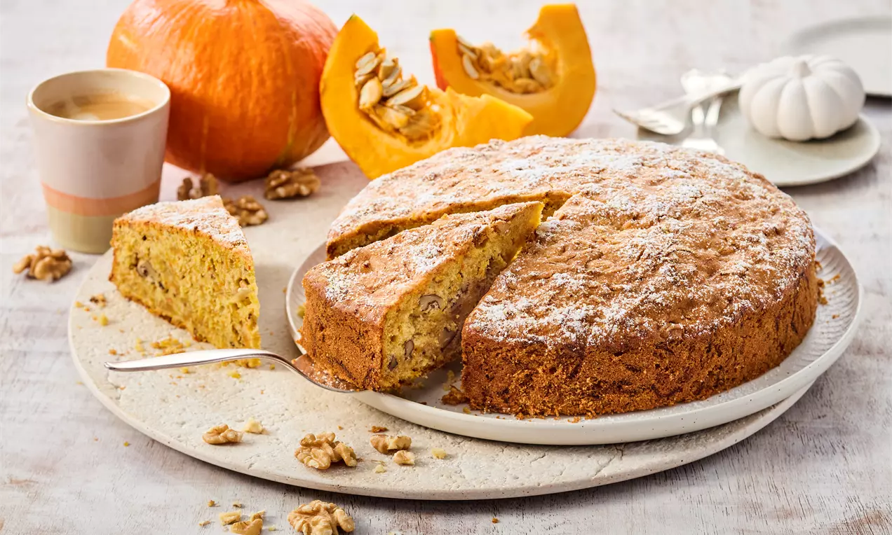 Saftiger Kürbiskuchen mit goldbrauner Kruste und zarter Struktur, angeschnitten auf einem Teller mit Walnüssen, Kürbisstücken und Kaffee