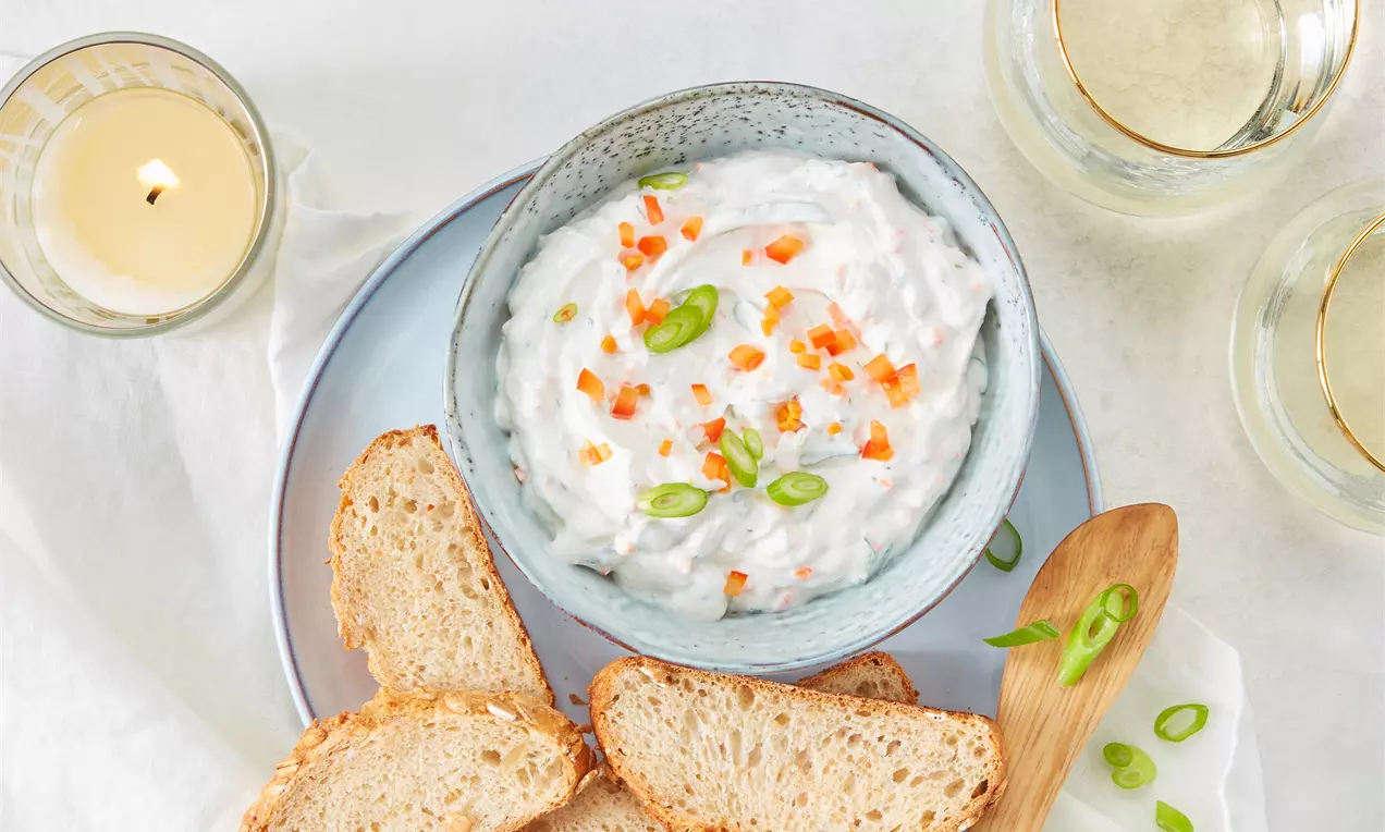 Cremiger weißer Dip in einer pastellblauen Schale, garniert mit orangefarbenen Würfeln und grünen Zwiebelringen, serviert mit Brotscheiben