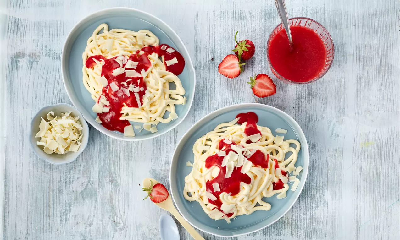 Spaghetti-Eis aus heller Vanillecreme mit roter Erdbeersoße und weißen Schokoraspeln auf blauen Tellern mit frischen Erdbeeren