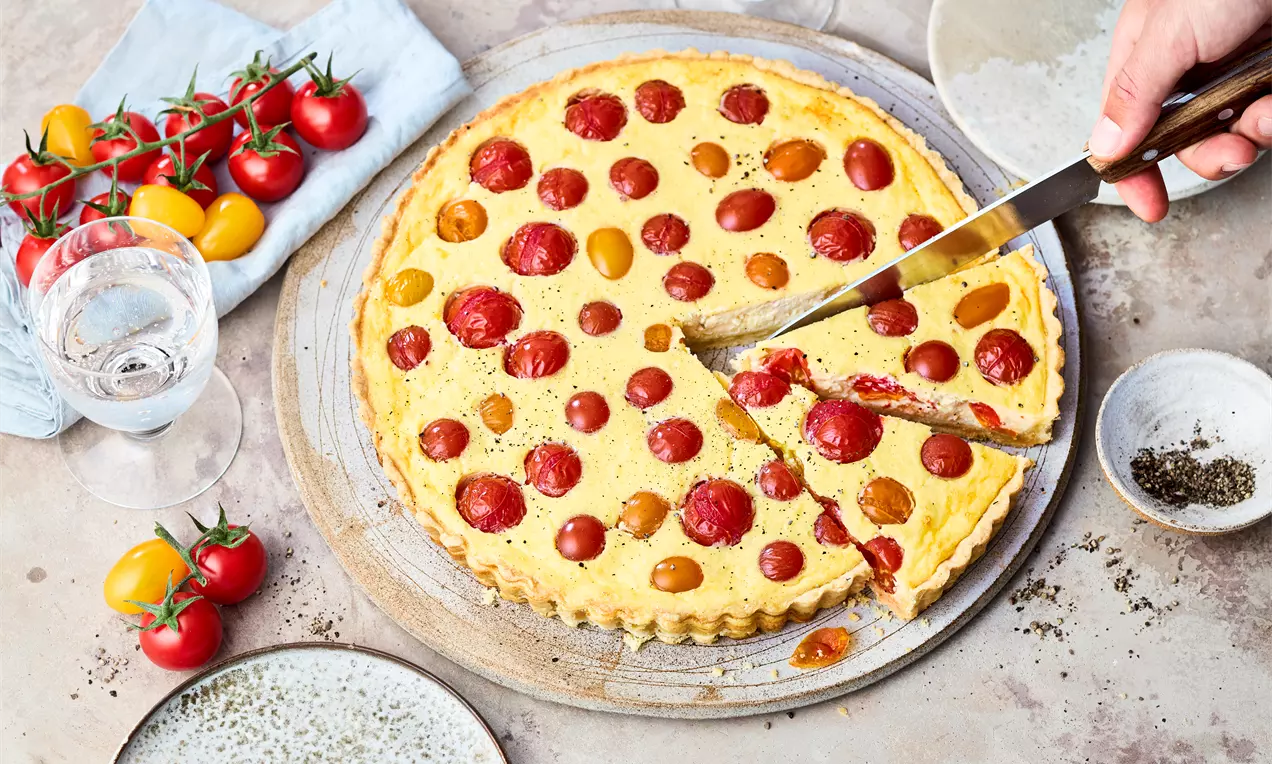 Goldgelbe Quiche mit roten und gelben Tomaten, auf einer hellen Holzplatte serviert, angeschnitten mit sichtbarer Füllung und umgeben von frischen Tomaten