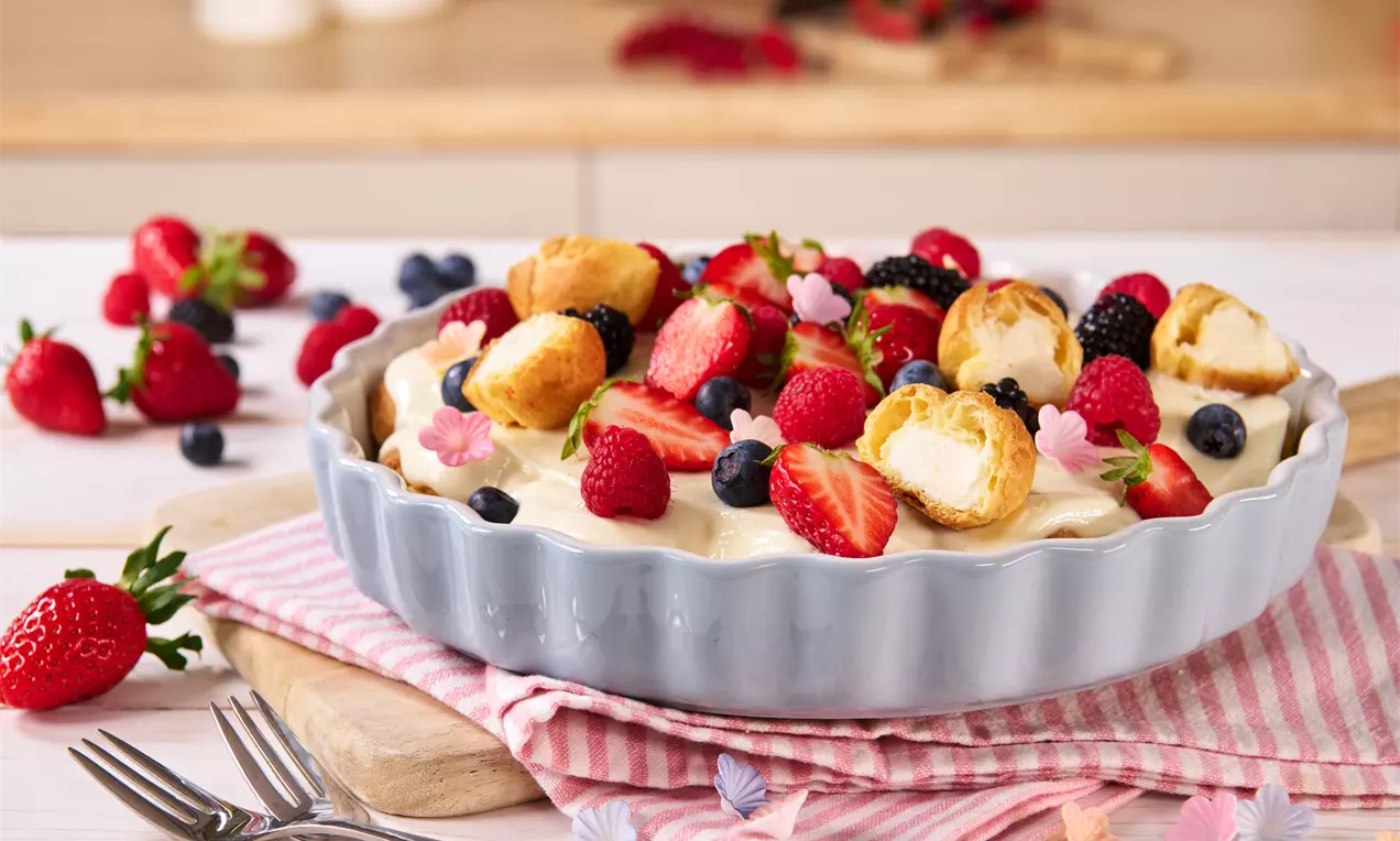 Ein Windbeutel-Dessert in einer cremefarbenen Schale, dekoriert mit frischen Beeren, essbaren Blüten und einer Sahnefüllung auf gestreiften Servietten