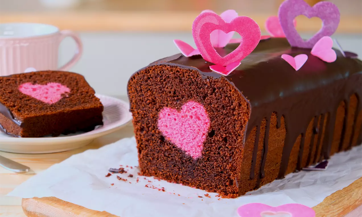 Saftiger Schokoladenkuchen mit rosa Herz und glänzend dunkler Glasur, dekoriert mit pinken und lila Herzformen auf weißem Papier