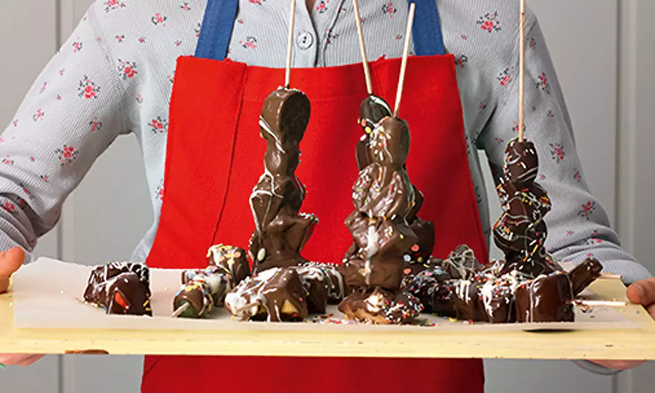 Schoko-Früchte-Spieße mit glänzender dunkler Glasur, bunten Streuseln, hoch aufgeschichtet auf Holzbrett, gehalten von Kind in roter Schürze