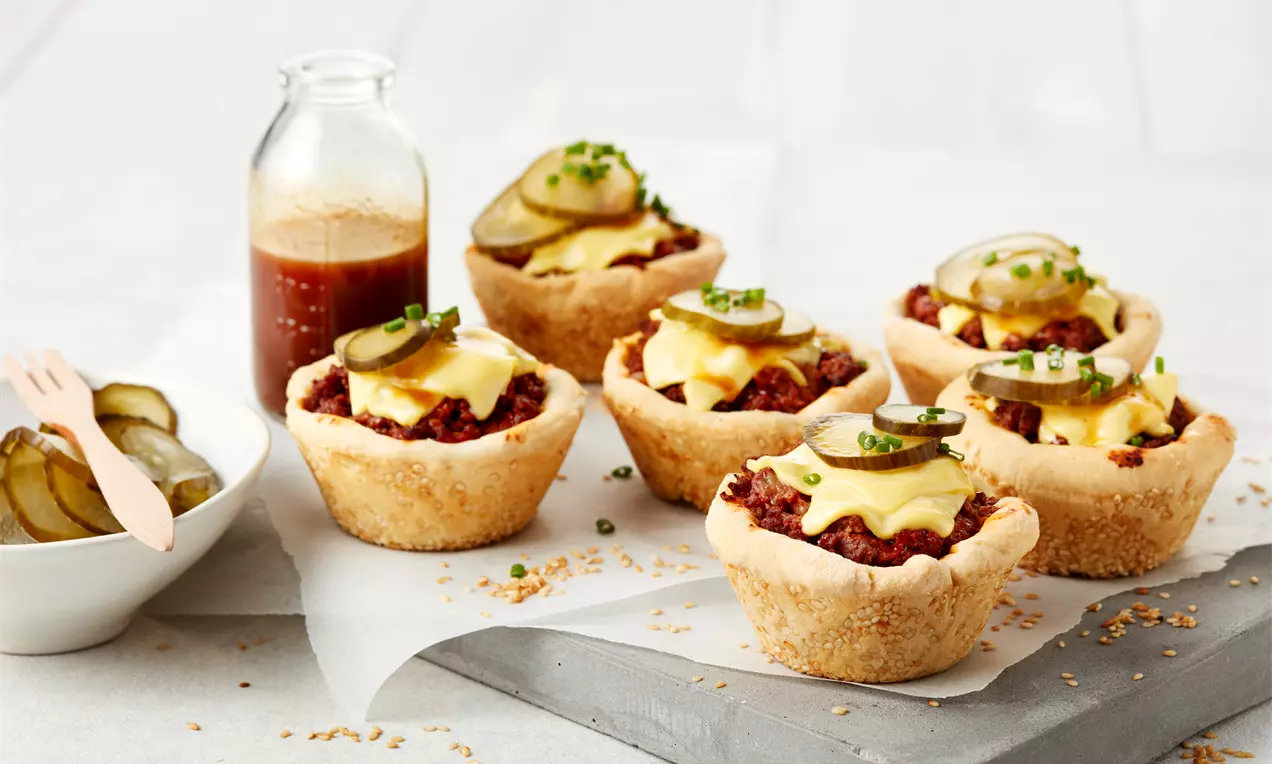 Sechs goldbraune Muffins aus knusprigem Teig mit saftigem Hackfleisch, geschmolzenem Käse, Gurkenscheiben und frischen Schnittlauchringen drapiert