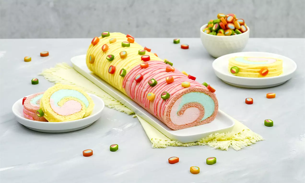 Bunte Biskuitrolle mit rosa, gelbem und blauem Wirbel, dekoriert mit farbenfrohen Fruchtgummi-Stücken auf weißer Platte mit Beilagen