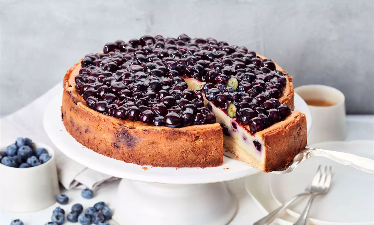 Käsekuchen mit goldbraunem Rand und glänzender Heidelbeer-Schicht, serviert auf einer weißen Tortenplatte mit angeschnittenem Stück