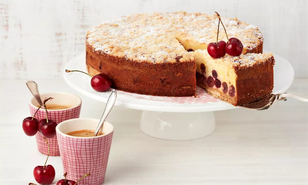 Rahm-Kirsch-Krümelkuchen mit goldbrauner Kruste, saftiger Kirschfüllung und Puderzucker auf weißer Tortenplatte, dekoriert mit frischen Kirschen