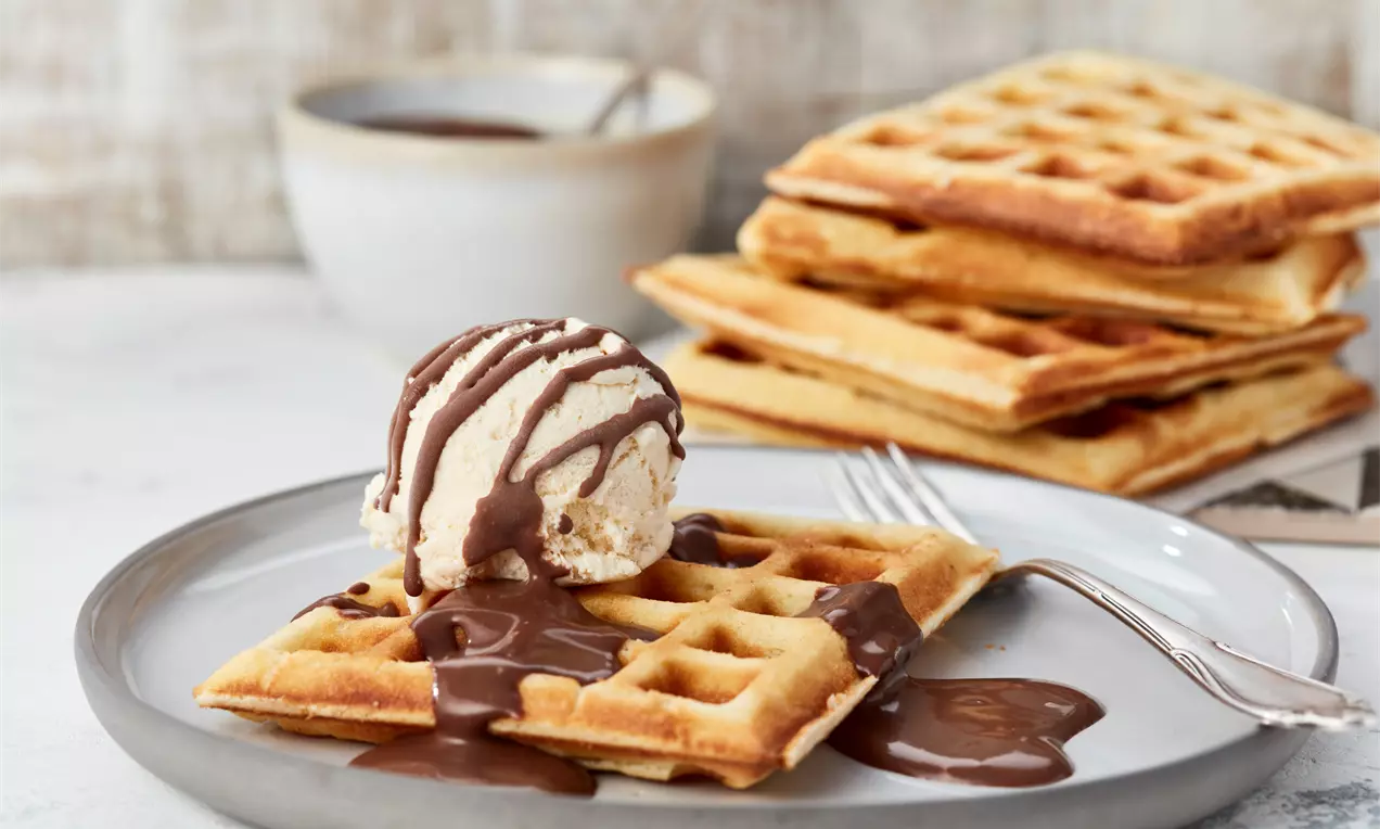 Goldene Brüsseler Waffel mit zartem Vanilleeis und glänzender Schokoladensauce auf grauem Teller, dahinter gestapelte Waffeln und Tasse