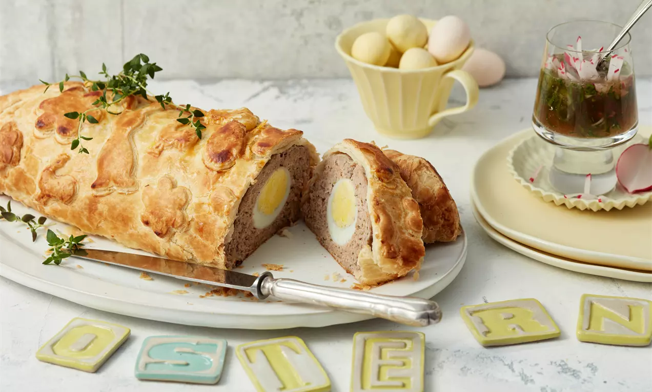 Ein goldbrauner Hackbraten im Blätterteigmantel mit dekorativem Ausstechmuster, angeschnitten und mit gekochtem Ei im Inneren, auf weißem Teller