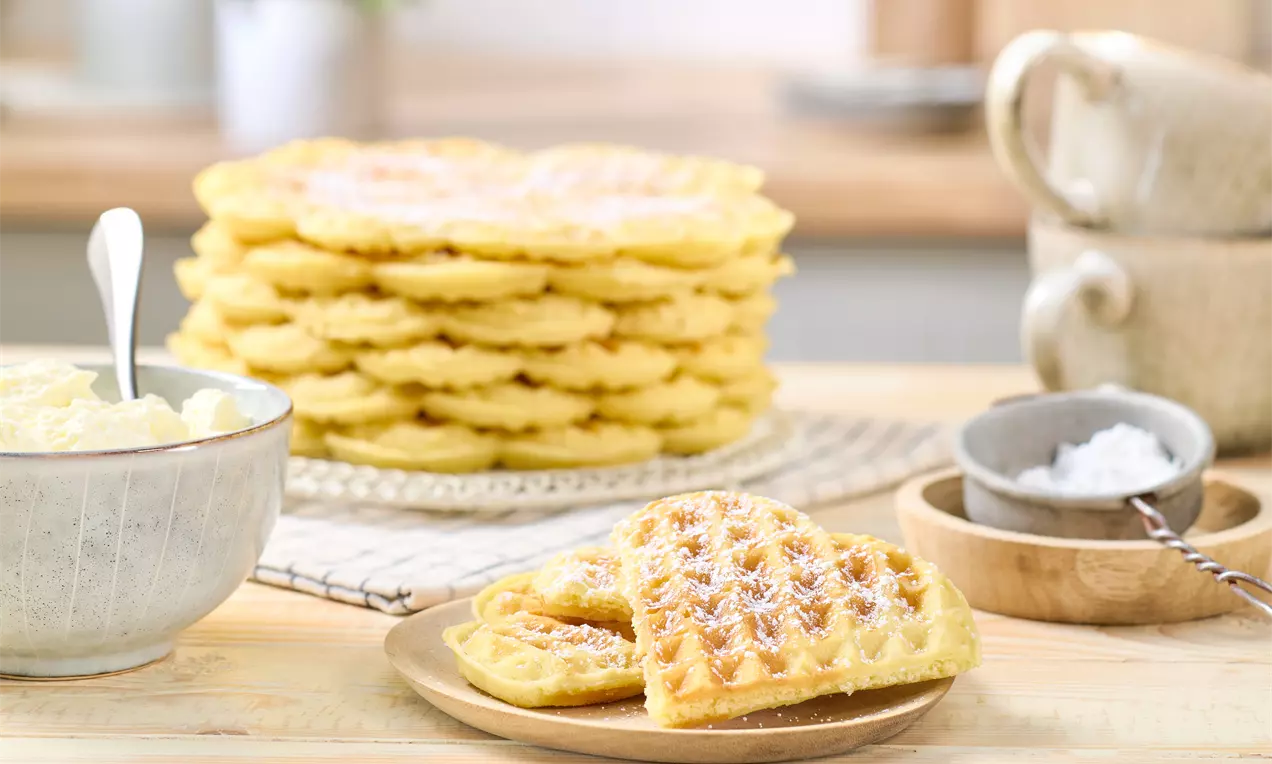 Gestapelte, goldbraune Waffeln auf einem Teller, im Vordergrund zwei Waffelstücke mit Puderzucker auf einem kleinen Teller. Daneben eine Schale mit Buttercreme und eine Schale mit Puderzucker