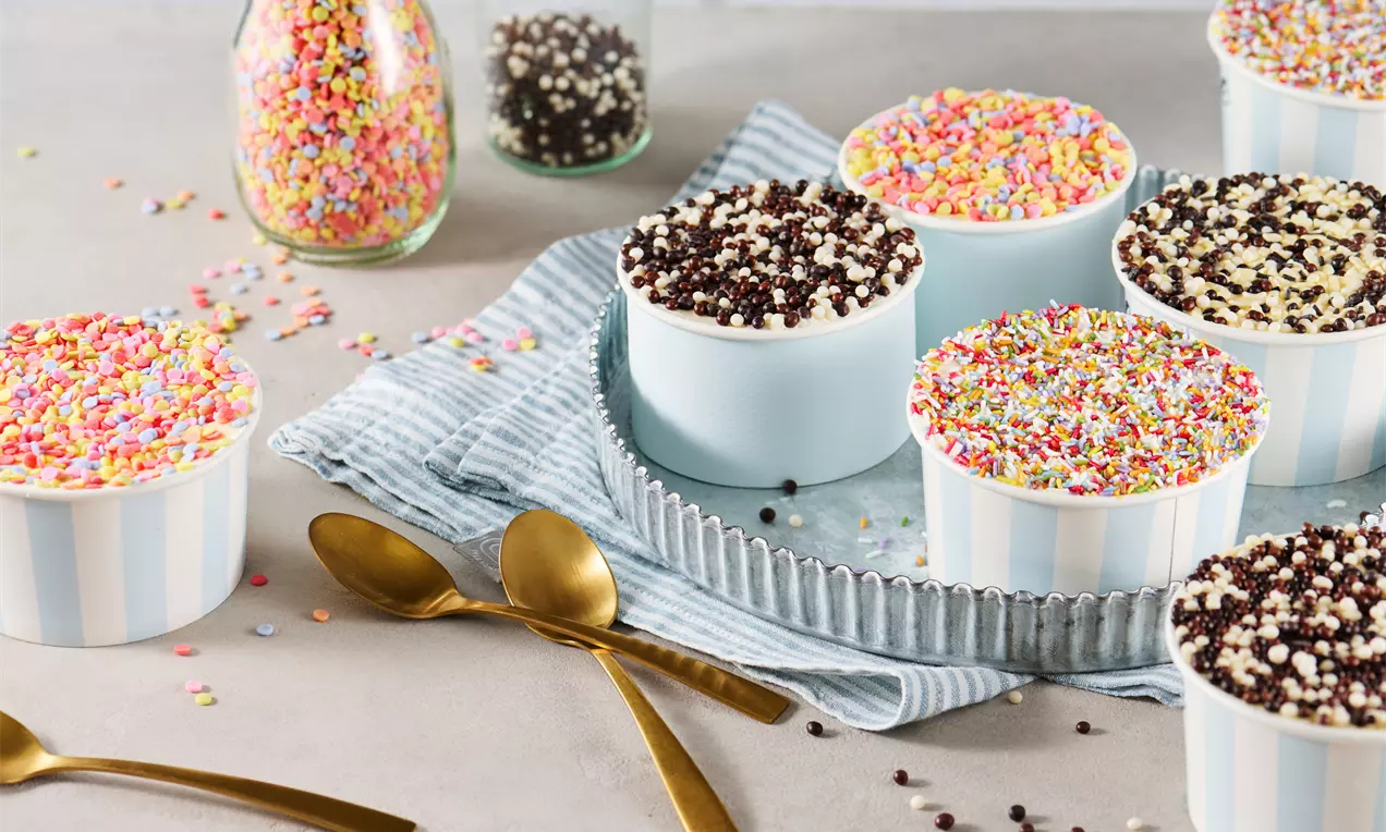 Pastellblaue Dessertschalen mit bunter Zuckerstreusel-Deko, auf einem grauen Tuch und Tablett arrangiert, umgeben von goldfarbenen Löffeln