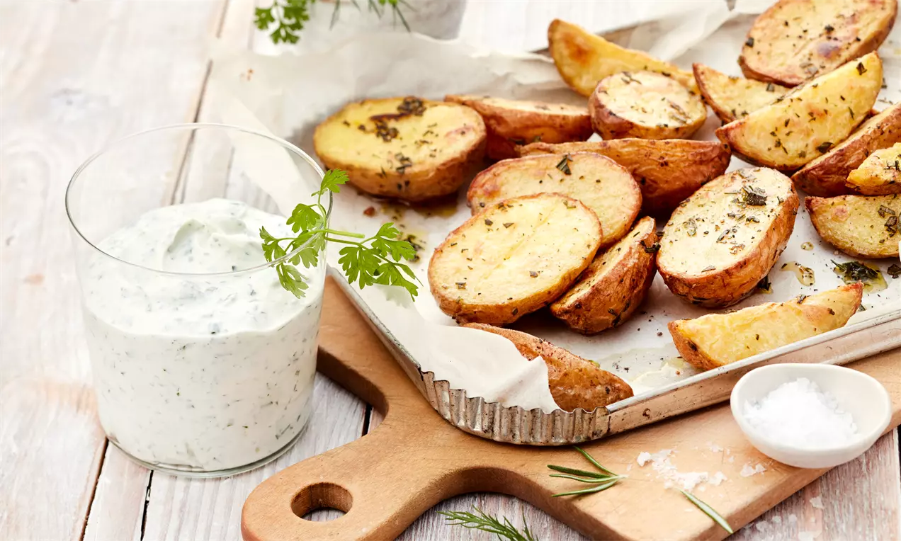 Goldene Kartoffelspalten mit Kräutern auf Backpapier neben einem Glas mit weißem Kräuterquark, garniert mit einem frischen Kräuterzweig