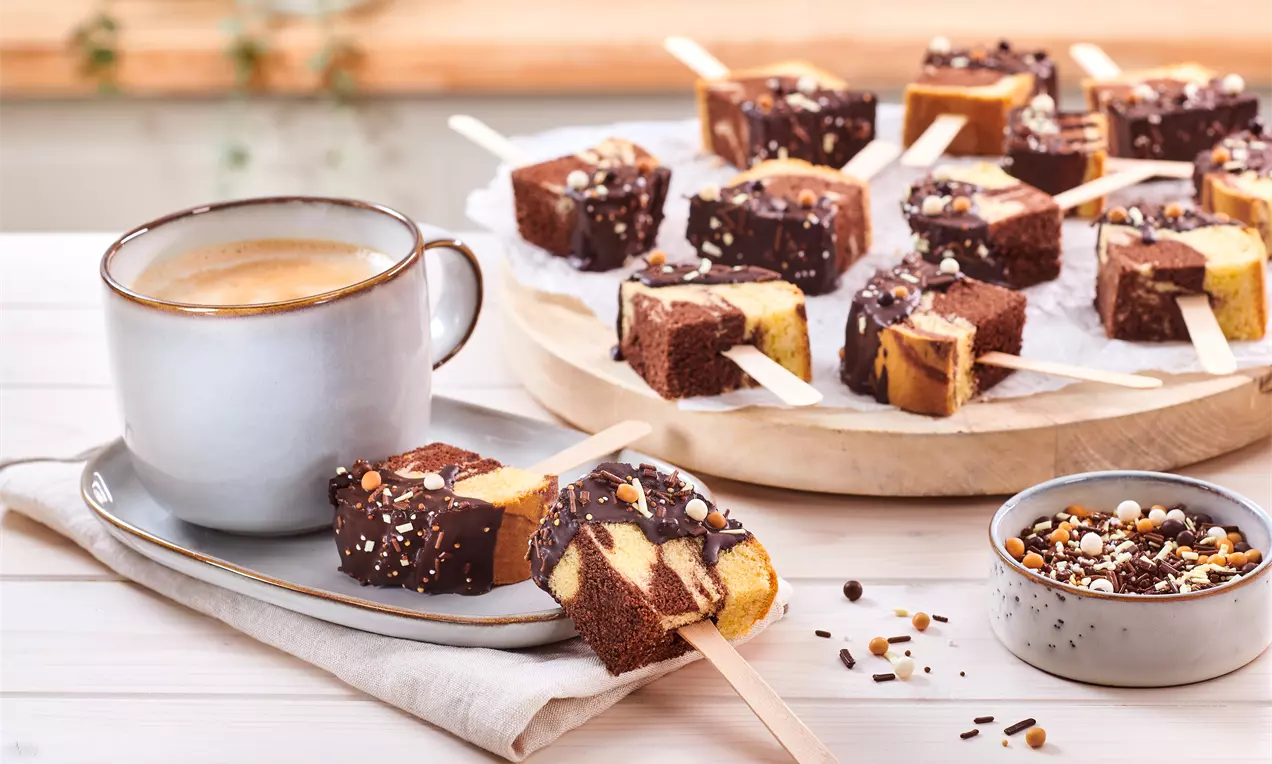 Marmorierte Cakesicles mit dunkler Schokoladenhülle und Streuseln, auf hellem Holztablett angerichtet, daneben Cappuccino und Schälchen
