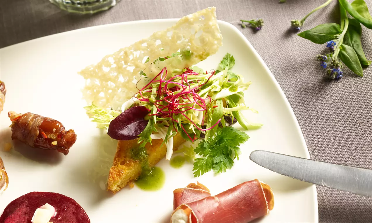 Maischnitten mit goldener Kruste, belegt mit frischem Kräutersalat, roten Sprossen und grüner Sauce auf einem weißen Teller mit Käsestange