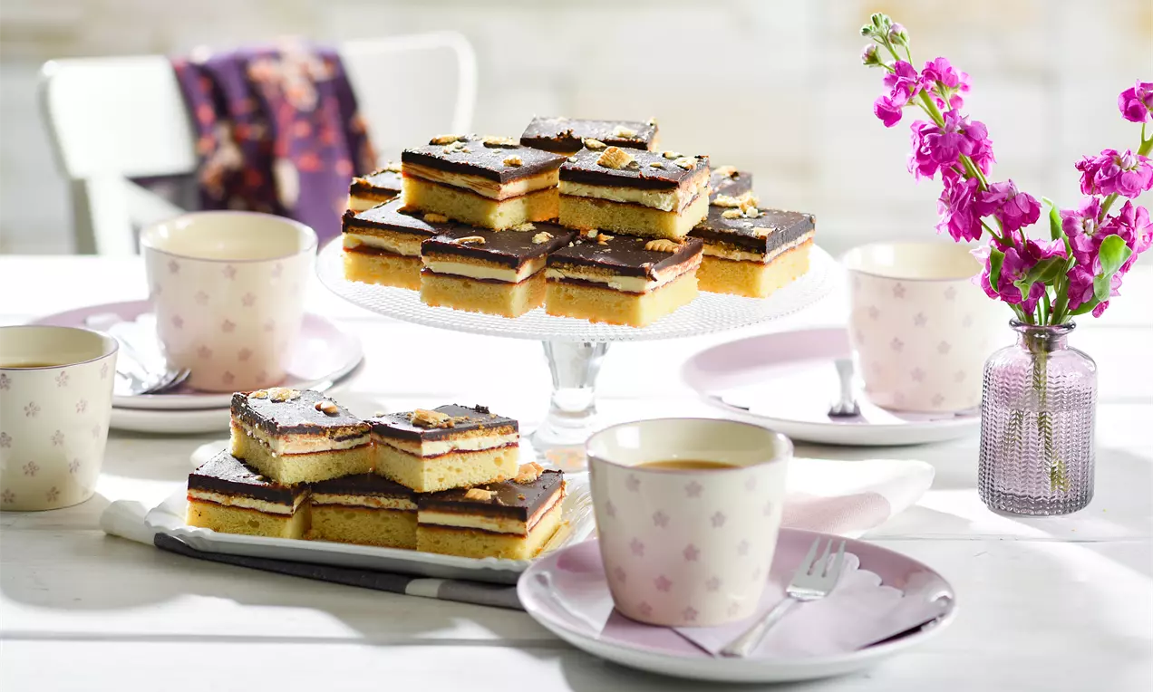 LPG-Kuchen mit goldgelbem Boden, dunkler Schokoladenschicht und Nusskrümeln auf Glasplatte, umgeben von blassrosa Geschirr und Blumenvasen