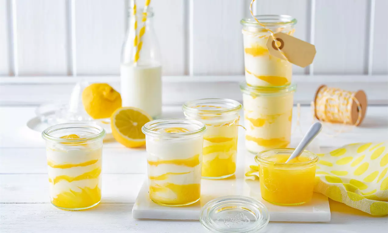 Weiße Mousse mit geschwungenen Gelb-Orangen-Lemon-Curd-Schichten in Gläsern, umgeben von Zitronen, Flasche und gelbem Tuch auf hellem Holz