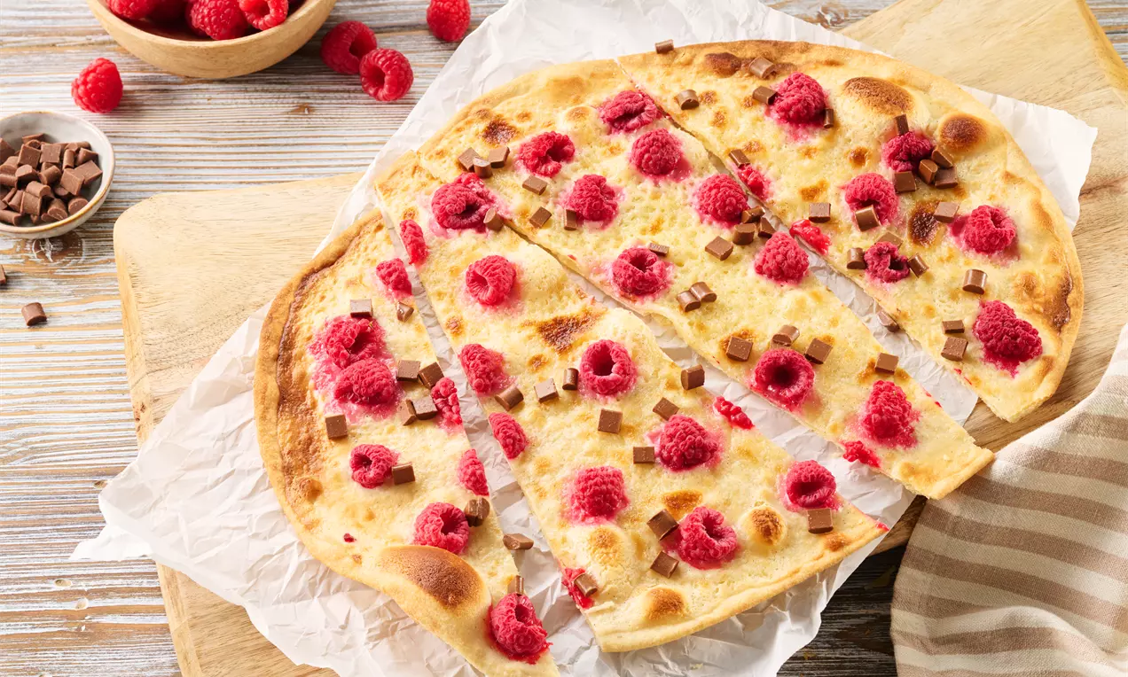 Dünner, goldbraun gebackener Flammkuchen mit frischen Himbeeren und kleinen Schokostückchen auf Backpapier auf einem Holzbrett serviert