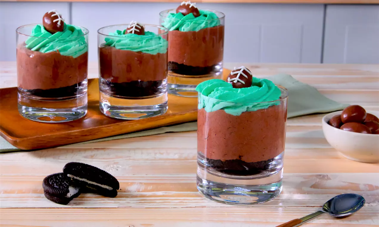 Schokocreme-Desserts im Glas mit Oreos, grüner gefärbter Sahne und kleinen Schoko-Bon-Footballs als Deko; vier Gläser auf Holztisch, dazu Kekse, Löffel und Schokokugeln.