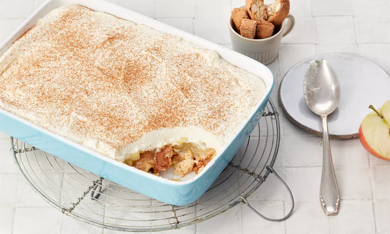 Apfel-Tiramisu in einer rechteckigen blauen Form, mit Schichten aus Sahne, Apfelstücken und Biscuits, bestreut mit Zimtpulver, daneben Löffel und Gebäck