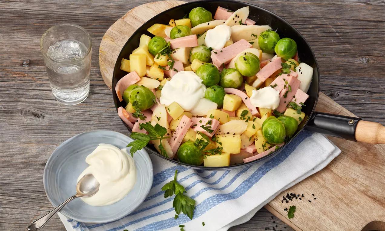 Bunte Pfanne mit goldgelben Kartoffelwürfeln, zarten Schwarzwurzelstücken, grünen Rosenkohlköpfen und rosa Schinkenstreifen mit Cremehaube