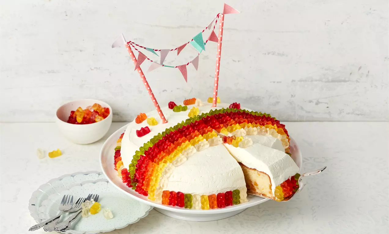 Bunter Gummibärchenkuchen mit Schichten von roten, gelben und grünen Gummibärchen auf Sahnehaube, angeschnitten auf weißem Teller mit Deko-Girlande