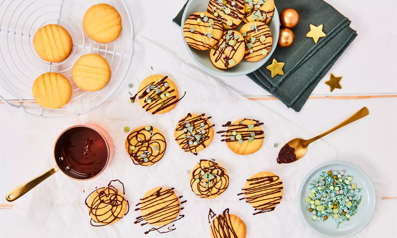 Runde goldgelbe Puddingplätzchen mit geschwungener Schokoglasur und bunten Streuseln auf weißem Papier neben Schüssel und Löffel drapiert