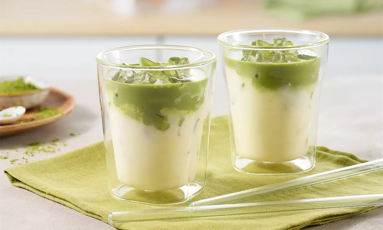 Zwei transparente Gläser mit geschichteter Iced Matcha Latte aus cremiger Milchbasis und leuchtend grüner Matcha-Schicht, serviert auf grünem Tuch.