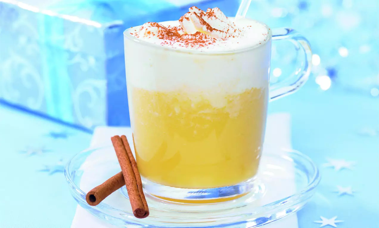 Weißwein-Orangen-Punsch in einer gläsernen Tasse mit cremigem Milchschaum, Zimtstreu und Zimtstangen auf hellem Untergrund serviert