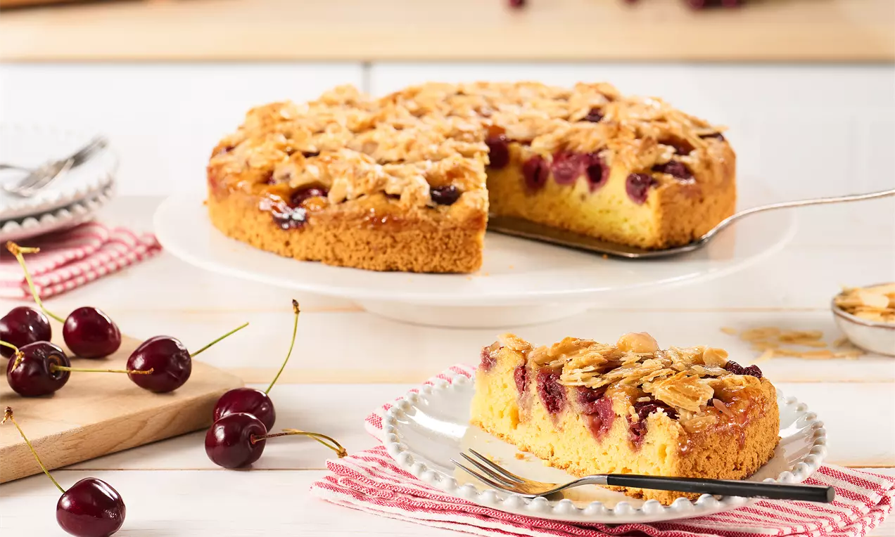 Goldbrauner Kirschkuchen mit süßen Kirschen und knusprigen Mandelblättchen auf weißer Tortenplatte, daneben ein angeschnittenes Stück