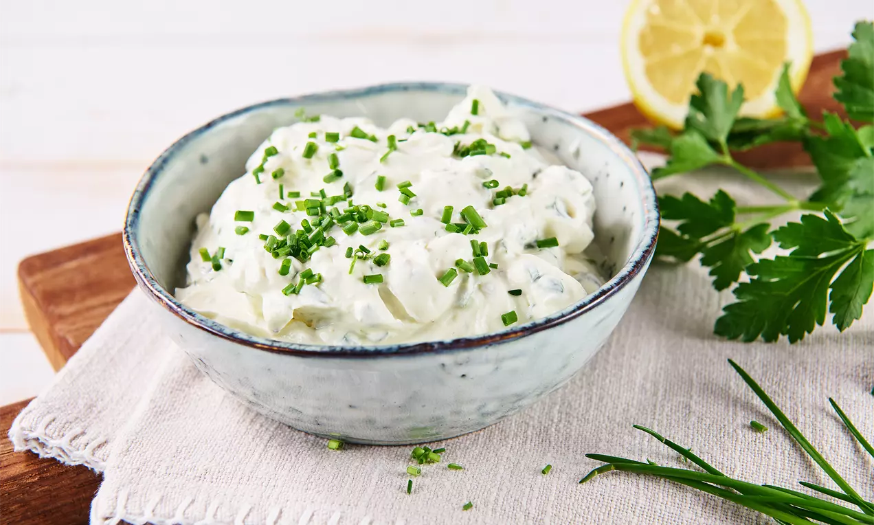 Cremiger Kräuterquark mit grünen Schnittlauchröllchen in einer weißen Keramikschale auf hellem Stofftuch, dekoriert mit frischen Kräutern und Zitrone