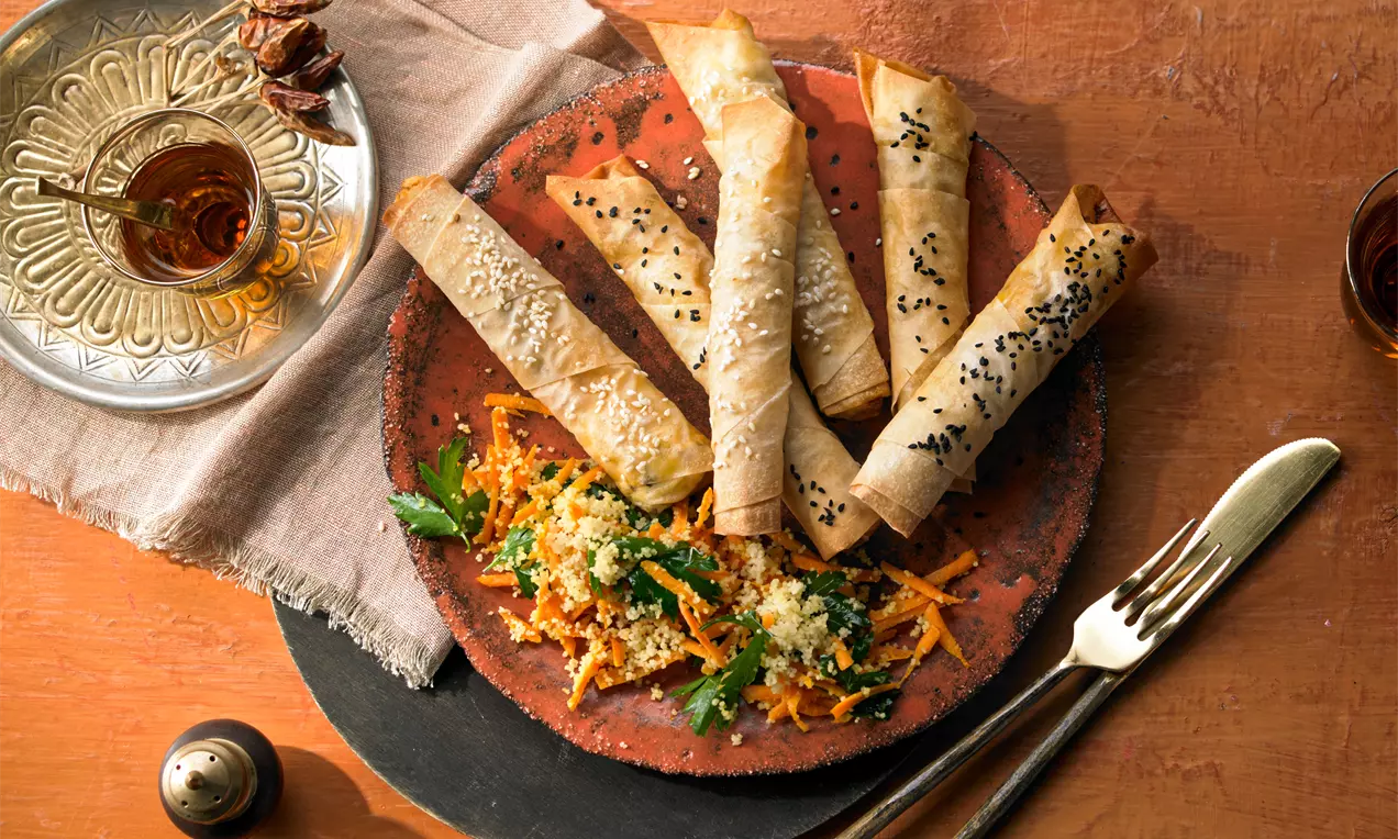 Knusprige Börek‑Cigar‑Rolls auf rustikalem Teller, teils mit Schwarzkümmel bestreut; serviert mit Möhren‑Kräuter‑Salat. Daneben Tee im verzierten Glas, Besteck und Leinentuch auf Holztisch.